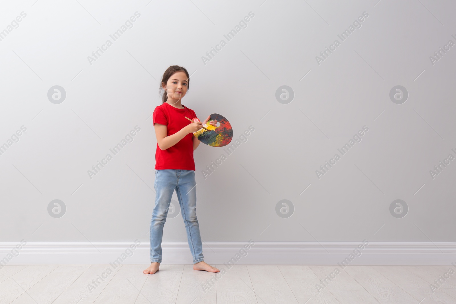 Photo of Little girl with brush and paint palette near light grey wall indoors. Space for text