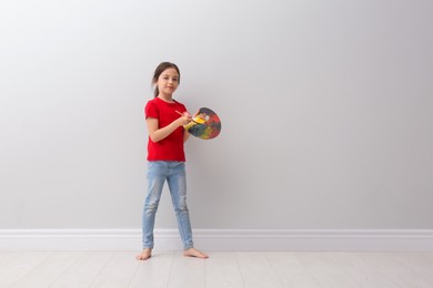 Little girl with brush and paint palette near light grey wall indoors. Space for text