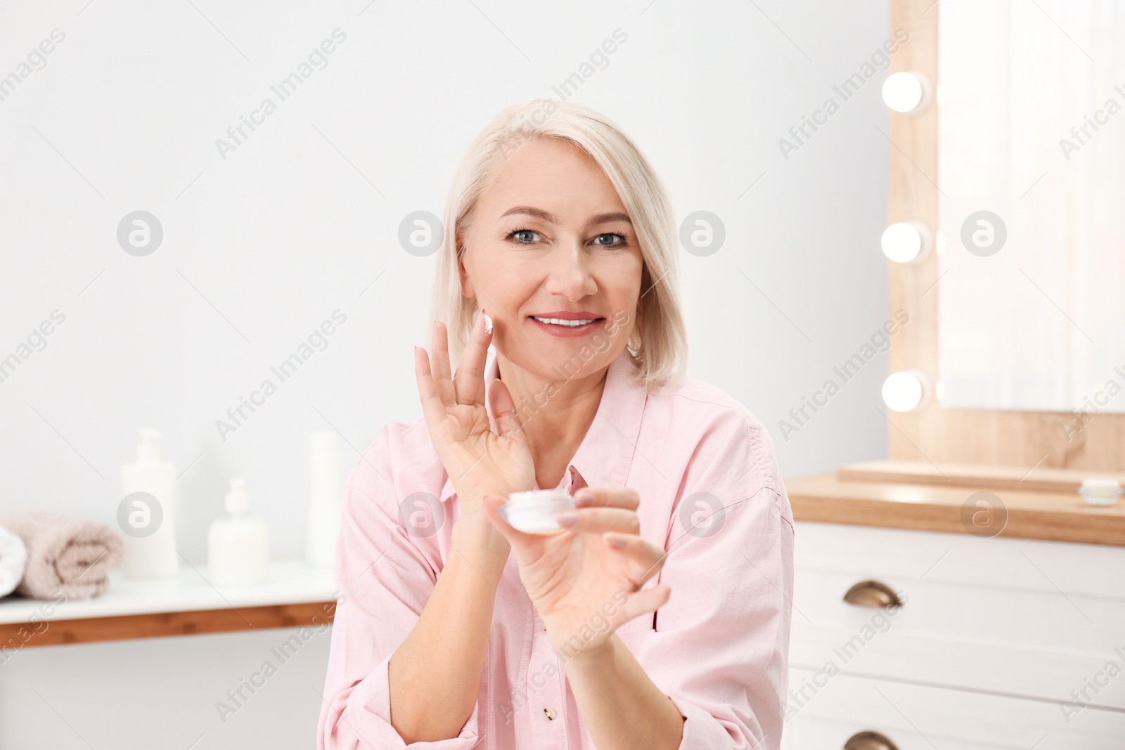 Photo of Portrait of charming mature woman with healthy beautiful face skin and natural makeup applying cream indoors