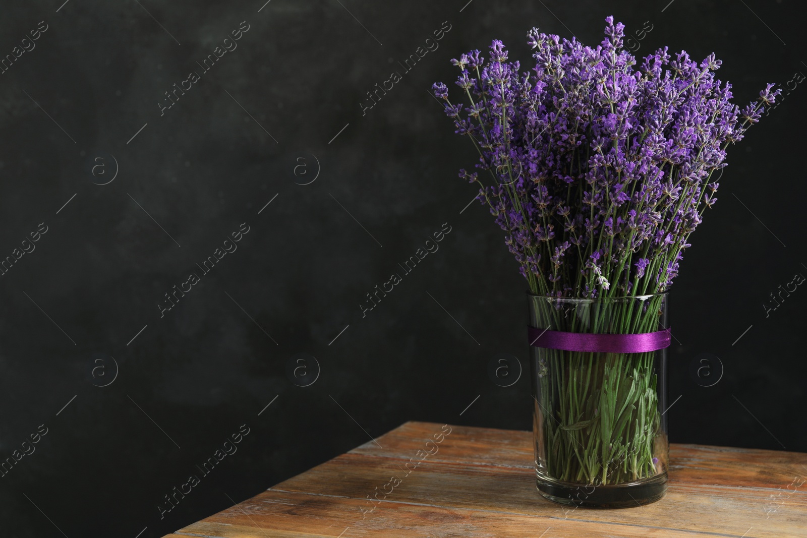 Photo of Beautiful lavender flowers in glass vase on wooden table against dark background. Space for text