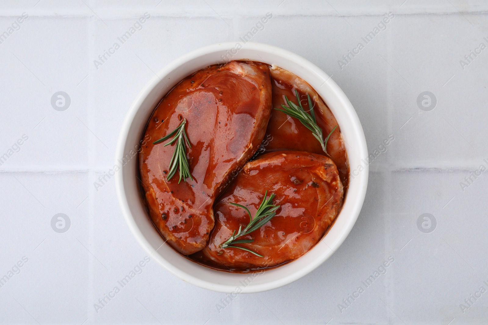 Photo of Raw marinated meat and rosemary in bowl on white tiled table, top view