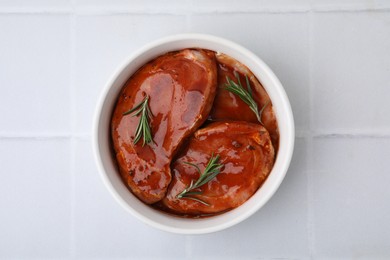 Raw marinated meat and rosemary in bowl on white tiled table, top view