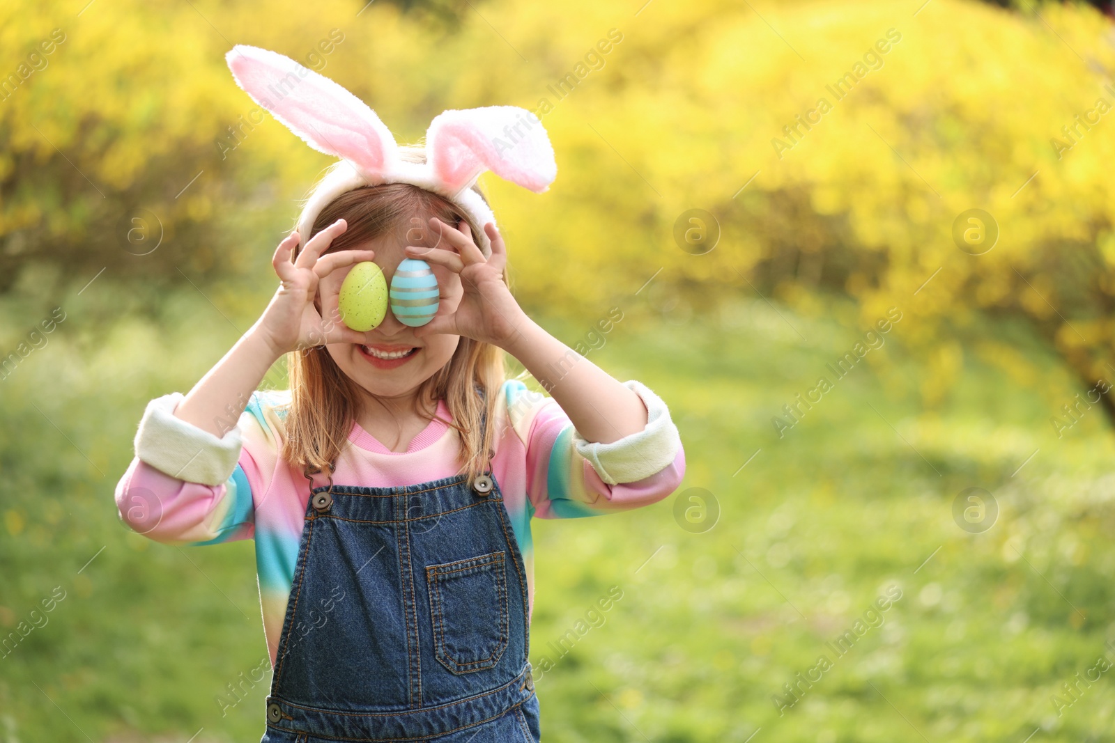 Photo of Easter celebration. Little girl in bunny ears covering eyes with painted eggs outdoors, space for text