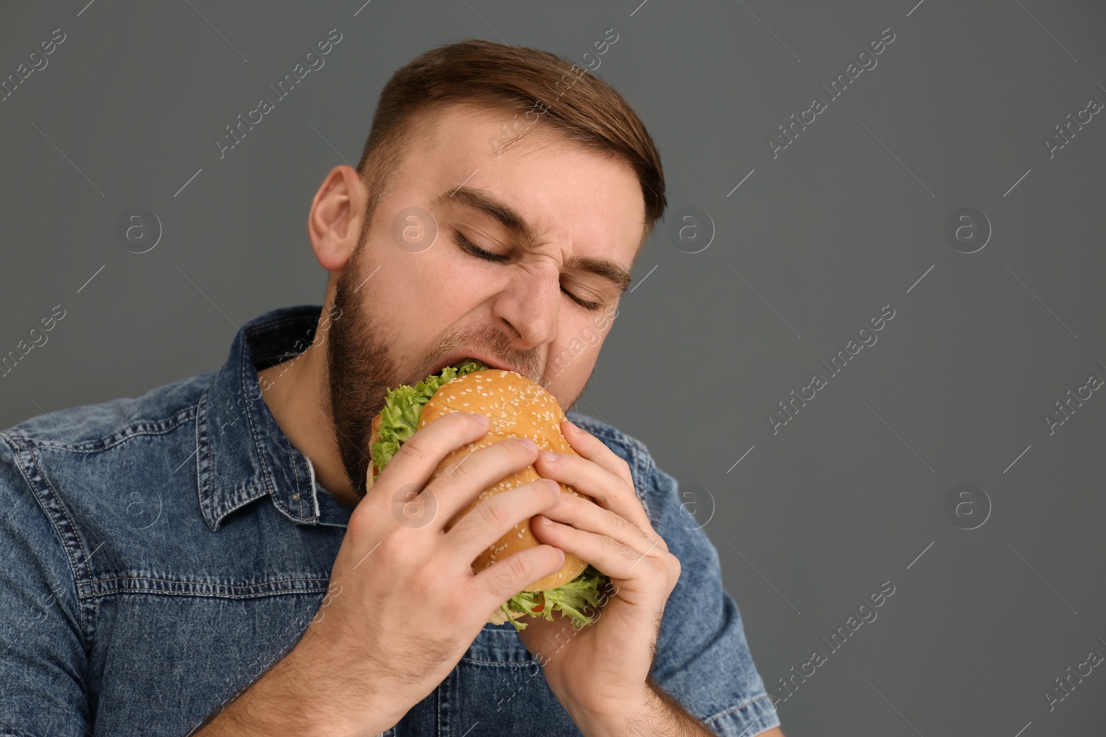 Photo of Young man eating tasty burger on grey background. Space for text