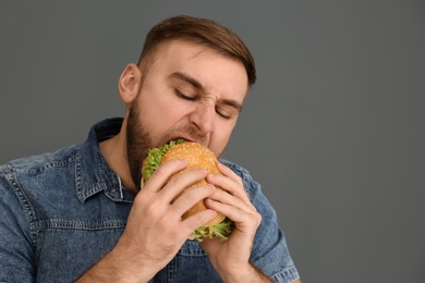 Young man eating tasty burger on grey background. Space for text