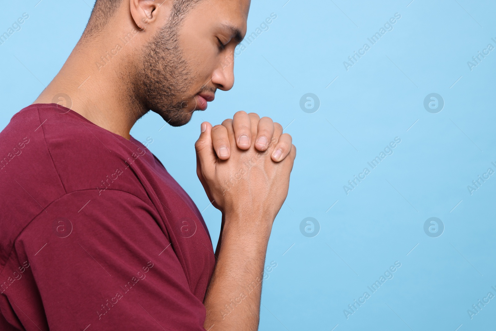 Photo of African American man with clasped hands praying to God on light blue background, closeup. Space for text