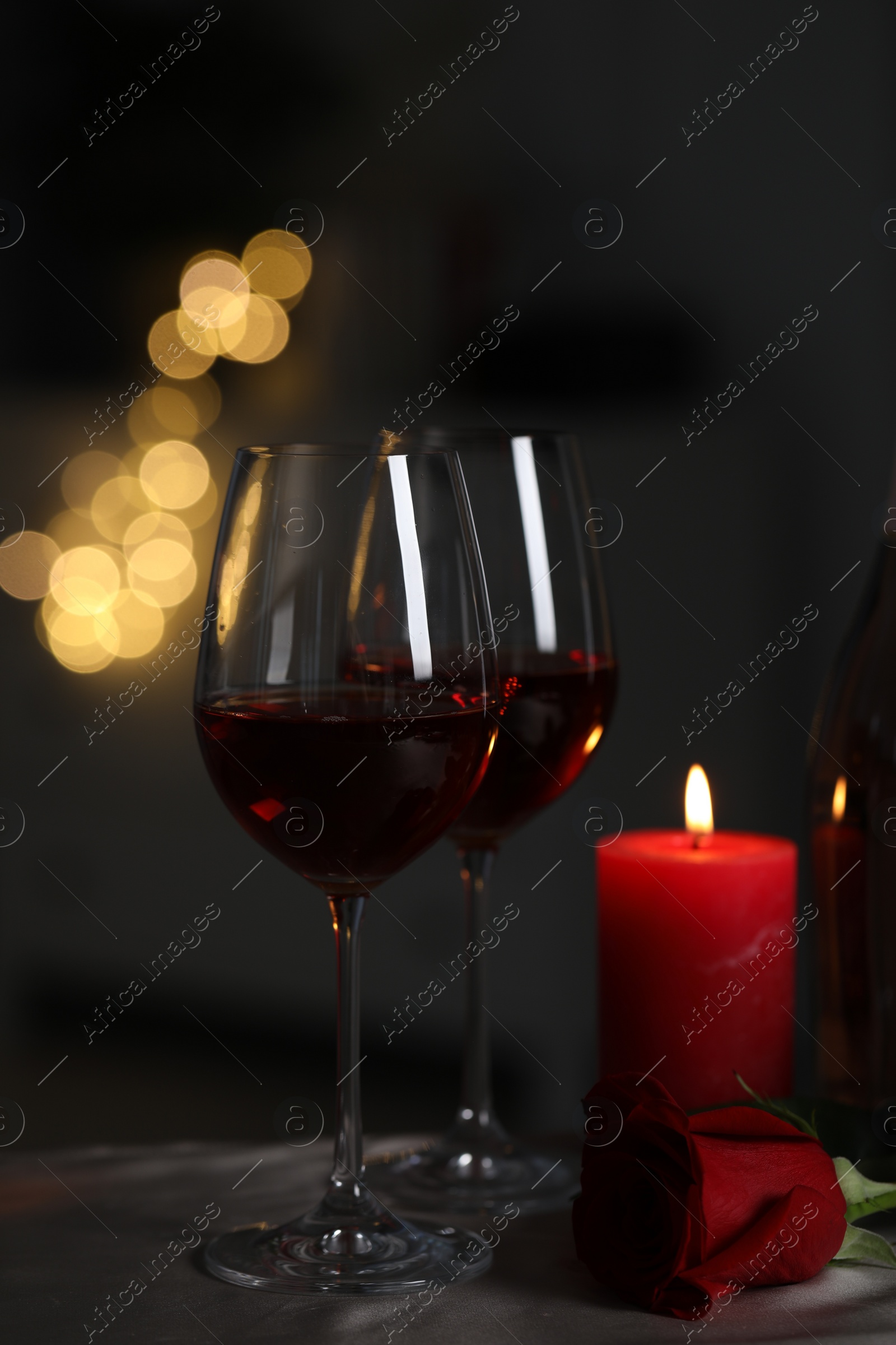 Photo of Glasses of red wine, rose flower and burning candle on grey table against blurred lights. Romantic atmosphere