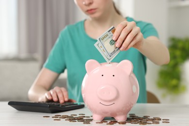 Financial savings. Woman putting dollar banknote into piggy bank at white wooden table indoors, closeup
