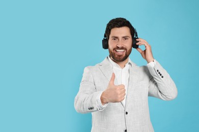Photo of Happy man listening music with headphones and showing thumb up on light blue background. Space for text