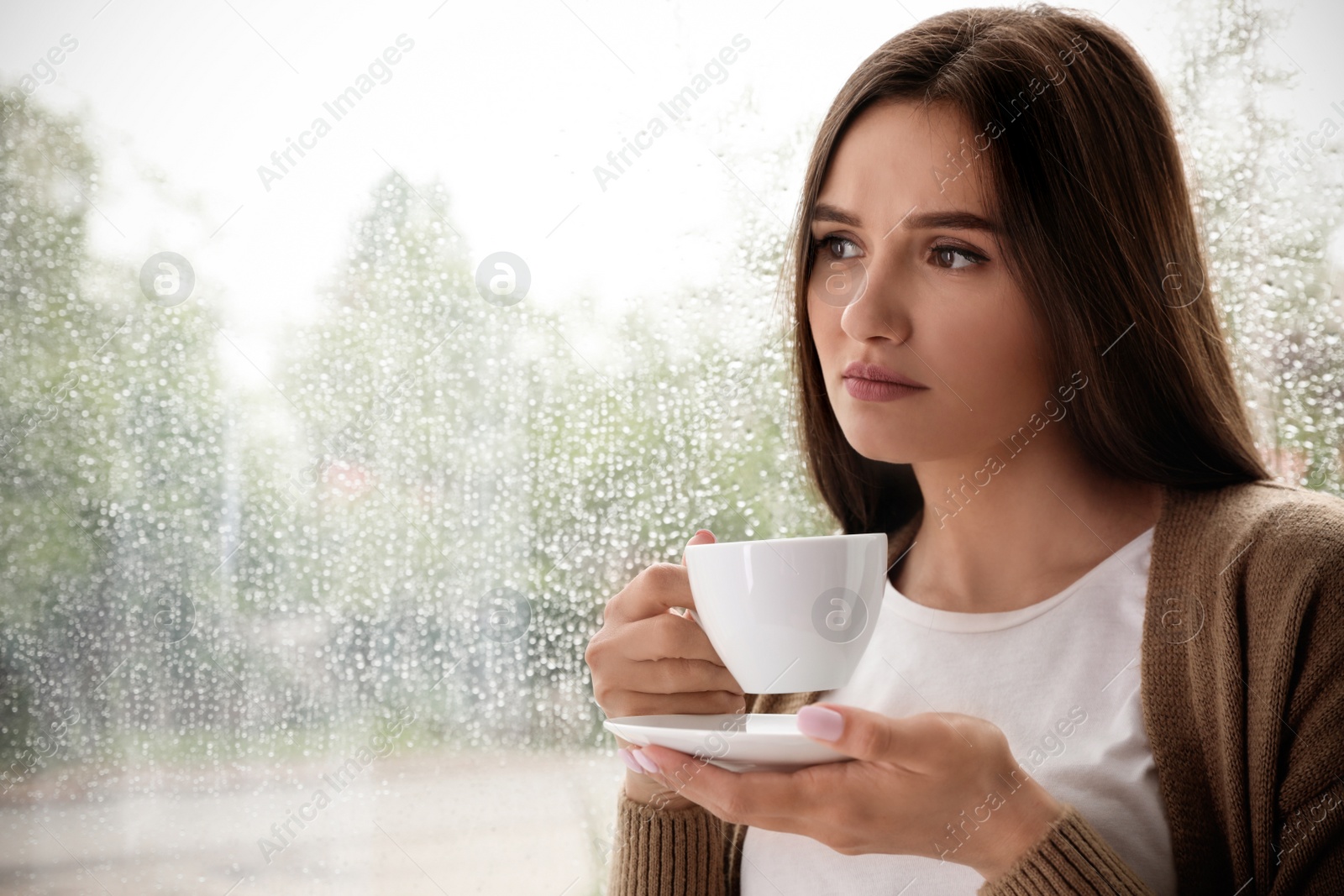 Photo of Thoughtful beautiful woman with cup of coffee near window indoors on rainy day