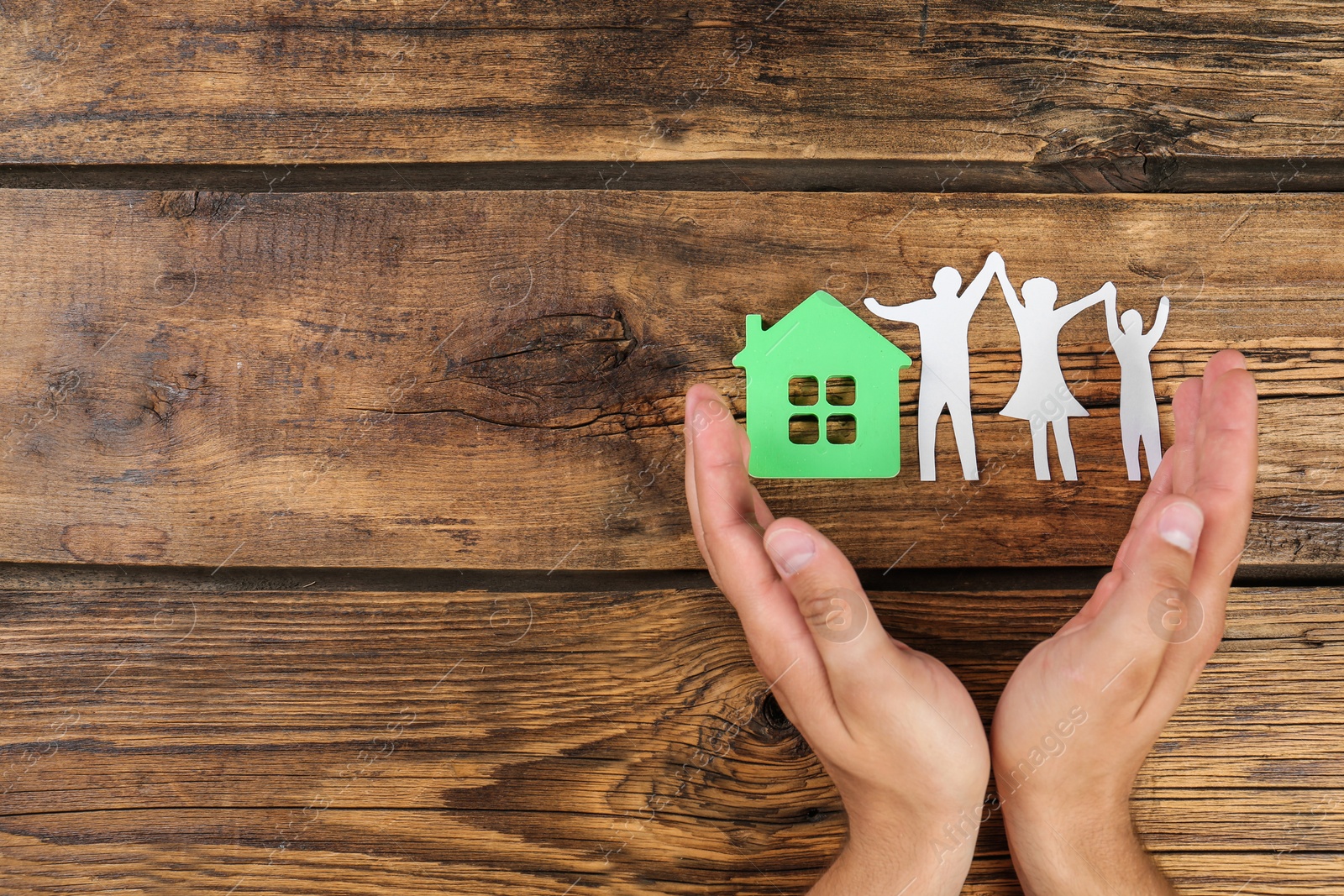 Photo of Young man protecting family figure and house model with his hands on wooden background, top view. Space for text