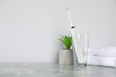 Photo of Electric toothbrush in glass, towels and green houseplant on light grey marble table near white wall. Space for text
