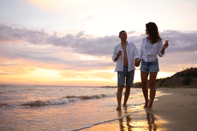 Photo of Lovely couple with glasses of wine walking on beach at sunset. Space for text