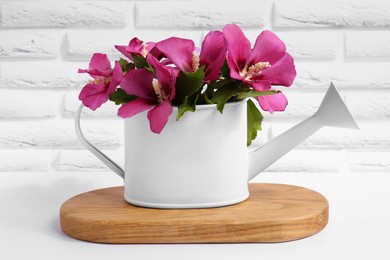 Beautiful flowers in watering can on white wooden table near brick wall