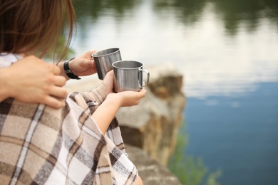 Photo of Cute couple with mugs and plaid near lake. Camping season