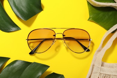 Photo of Stylish sunglasses and green leaves on yellow background, flat lay