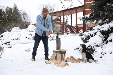 Photo of Man chopping wood with axe next to cute dog outdoors on winter day