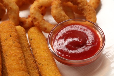 Photo of Tasty ketchup, cheese sticks and onion rings on white table, closeup