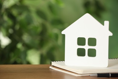 Photo of Mortgage concept. House model, notebook and pen on wooden table against blurred background, space for text