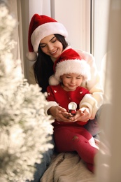 Photo of Mother and daughter in Santa hats playing with snow globe near window