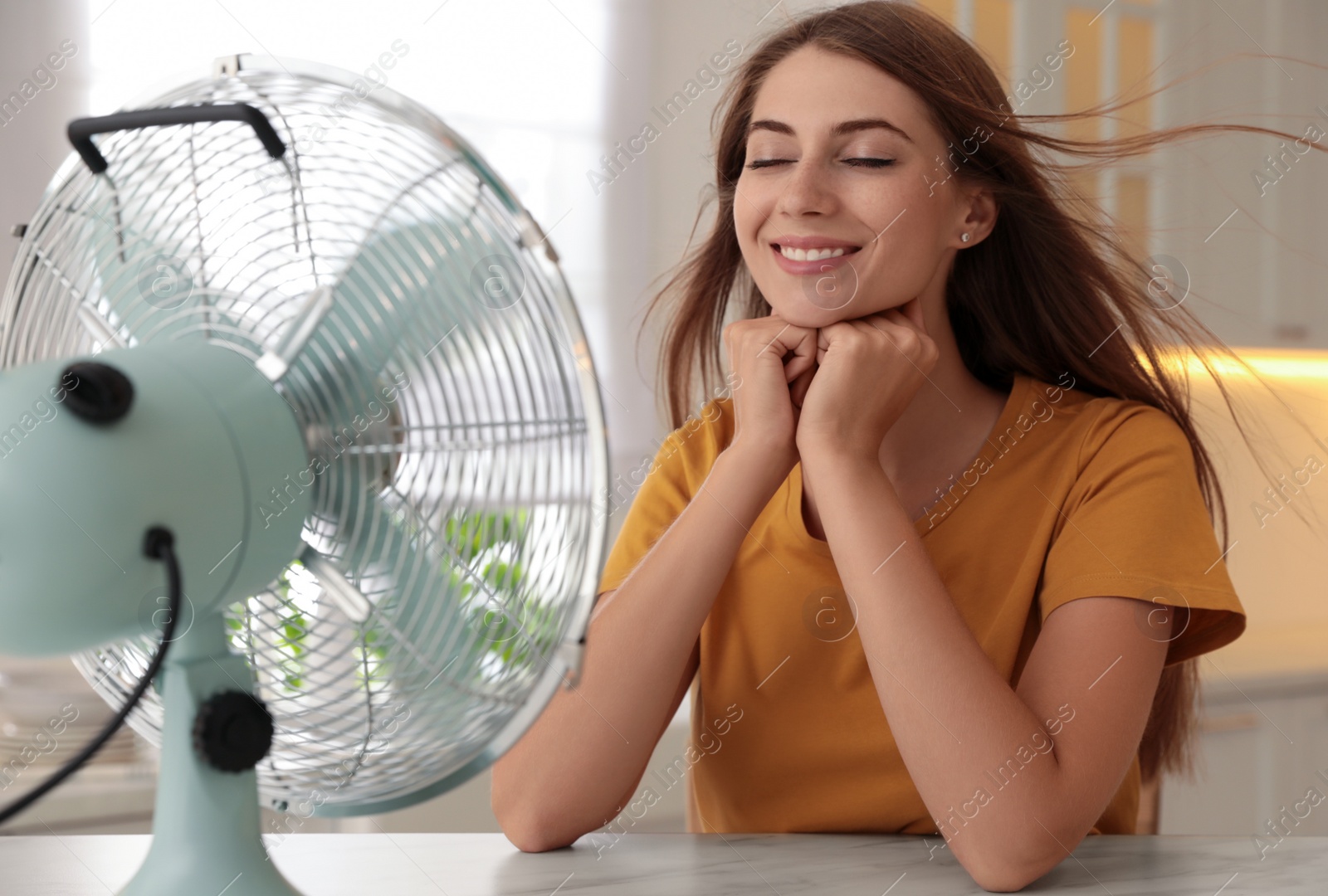 Photo of Woman enjoying air flow from fan at table in kitchen. Summer heat