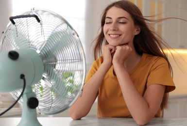 Photo of Woman enjoying air flow from fan at table in kitchen. Summer heat