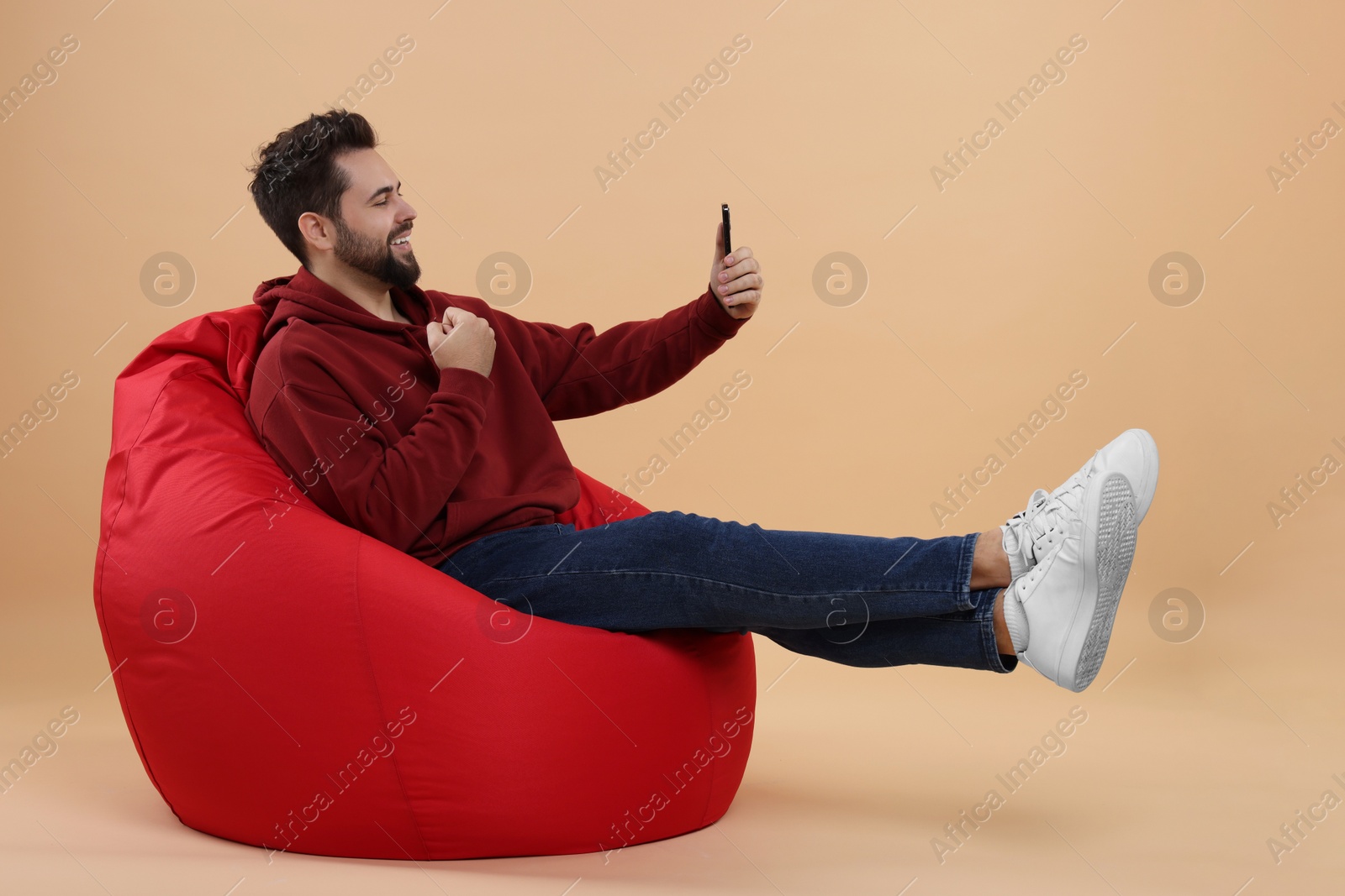 Photo of Happy young man using smartphone on bean bag chair against beige background