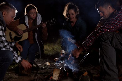 Photo of Group of friends roasting marshmallows on bonfire at camping site in evening