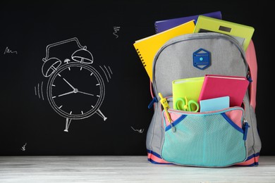 Image of Backpack full of stationery near blackboard with drawn alarm clock. Time to school