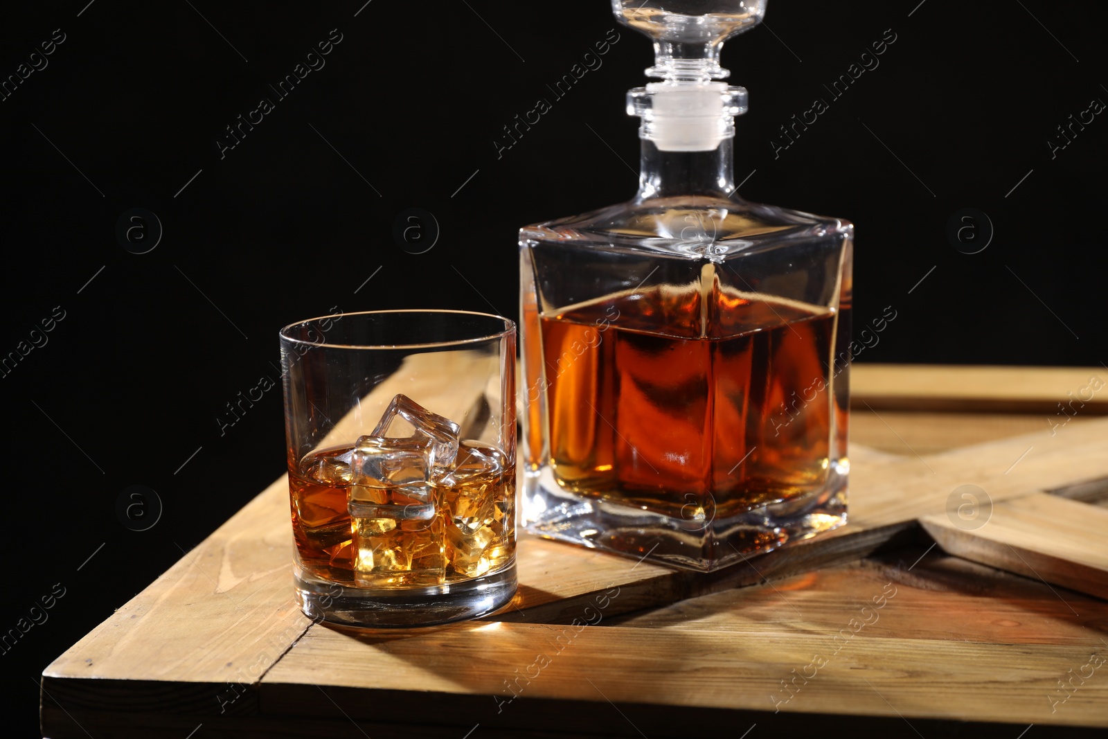 Photo of Whiskey with ice cubes in glass and bottle on wooden crate against black background, closeup