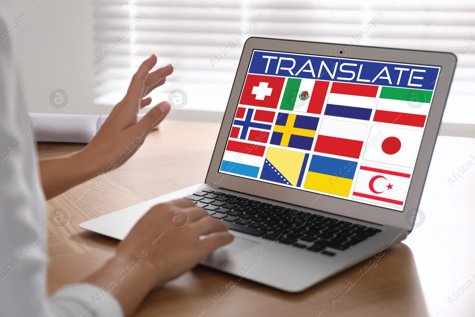 Image of Translator using modern laptop with images of different flags on screen at wooden table indoors, closeup