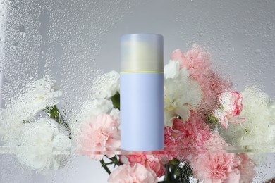 Bottle with moisturizing cream and beautiful flowers on light background, view through wet glass