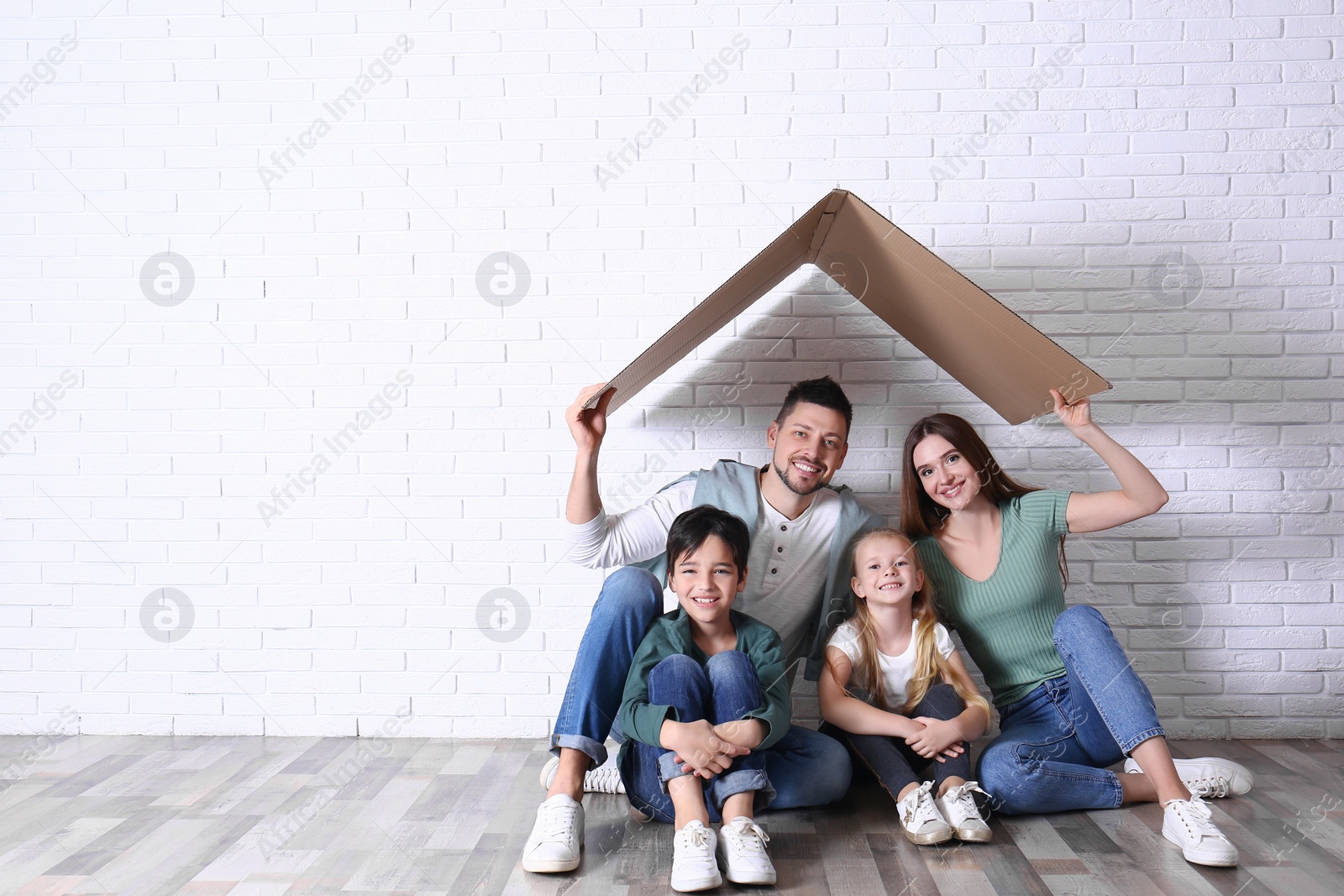 Photo of Happy family sitting under cardboard roof near brick wall, space for text. Insurance concept