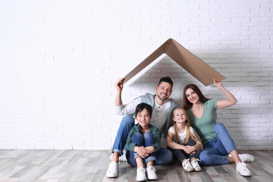 Happy family sitting under cardboard roof near brick wall, space for text. Insurance concept