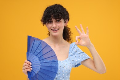 Photo of Happy woman holding hand fan and showing ok gesture on orange background