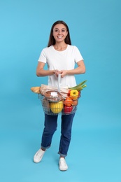 Photo of Young woman with shopping basket full of products on blue background