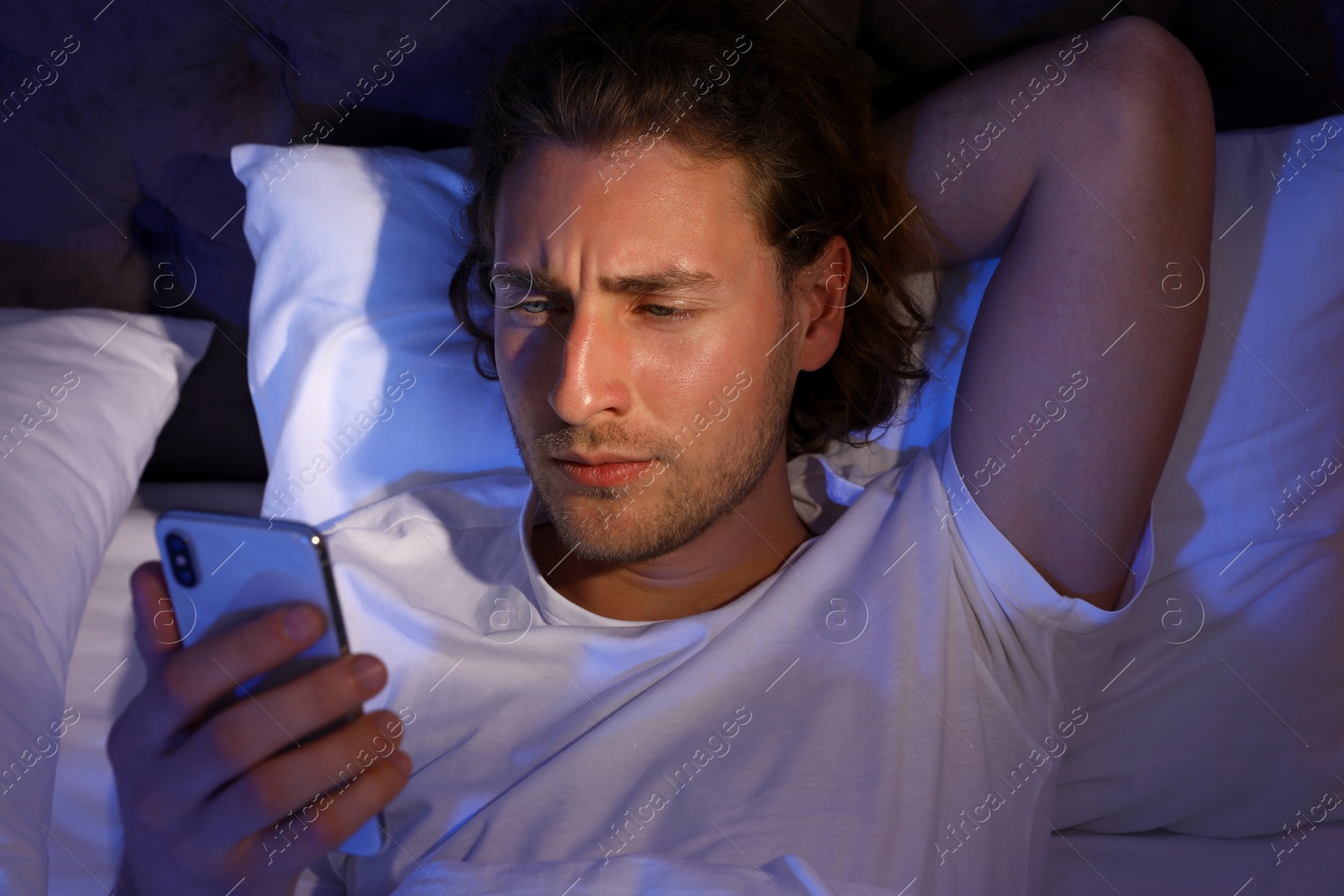 Photo of Handsome young man using smartphone while lying on pillow at night, view from above. Bedtime