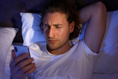 Handsome young man using smartphone while lying on pillow at night, view from above. Bedtime