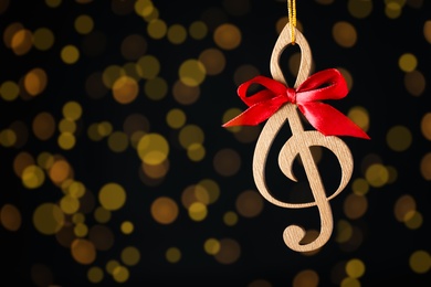 Wooden music note with red bow hanging on black background with blurred Christmas lights. Space for text