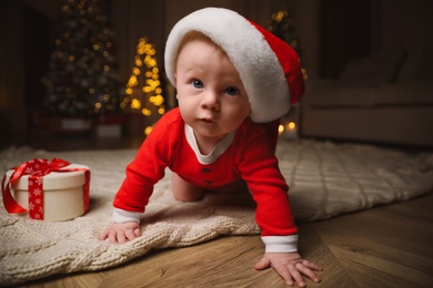 Cute baby in Santa hat with Christmas gift on floor at home