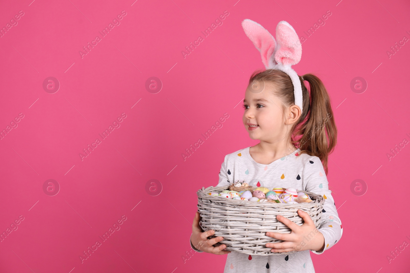 Photo of Adorable little girl with bunny ears holding wicker basket full of Easter eggs on pink background. Space for text