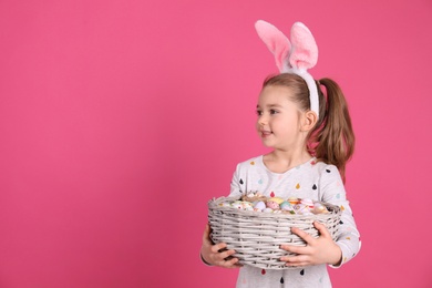 Adorable little girl with bunny ears holding wicker basket full of Easter eggs on pink background. Space for text