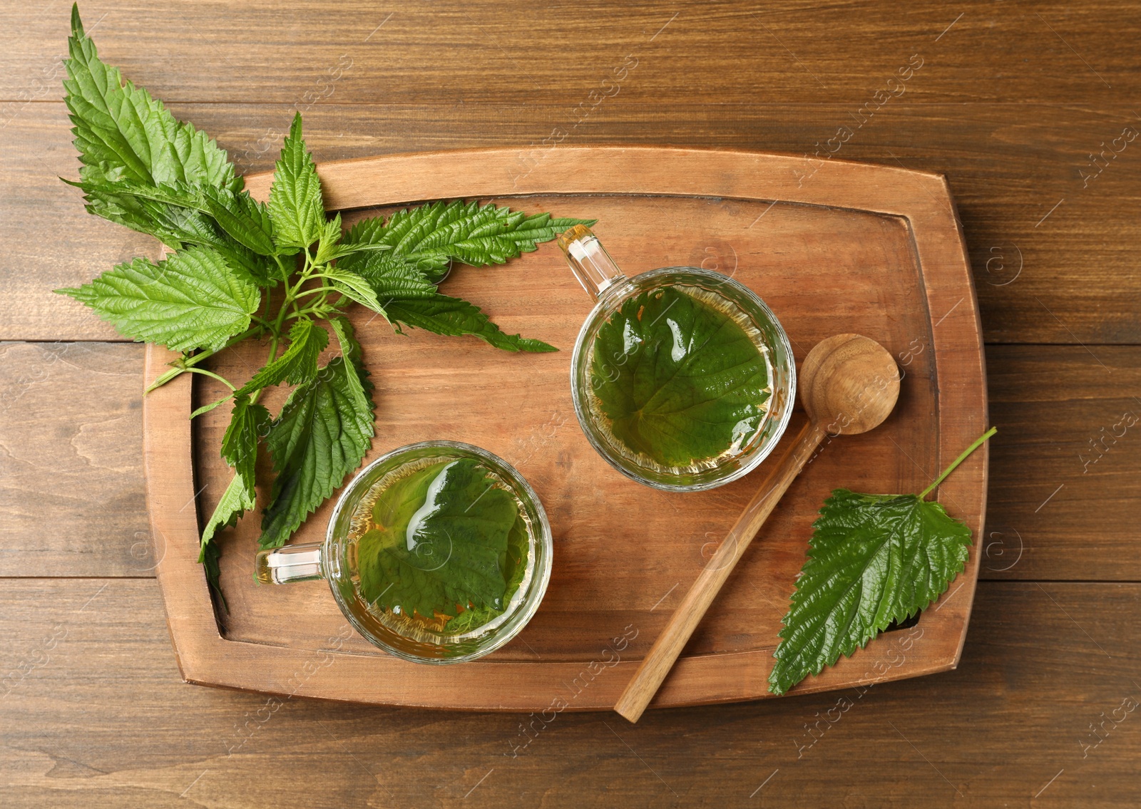 Photo of Glass cups of aromatic nettle tea and green leaves on wooden table, top view