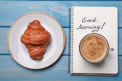 Aromatic coffee, croissant and Good Morning! message on light blue wooden table, flat lay