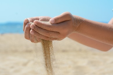 Photo of Child pouring sand from hands on beach near sea, closeup. Fleeting time concept