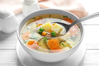 Spoon of fresh homemade vegetable soup over full bowl on white wooden table, closeup