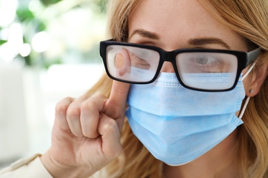 Photo of Woman wiping foggy glasses caused by wearing medical mask indoors, closeup