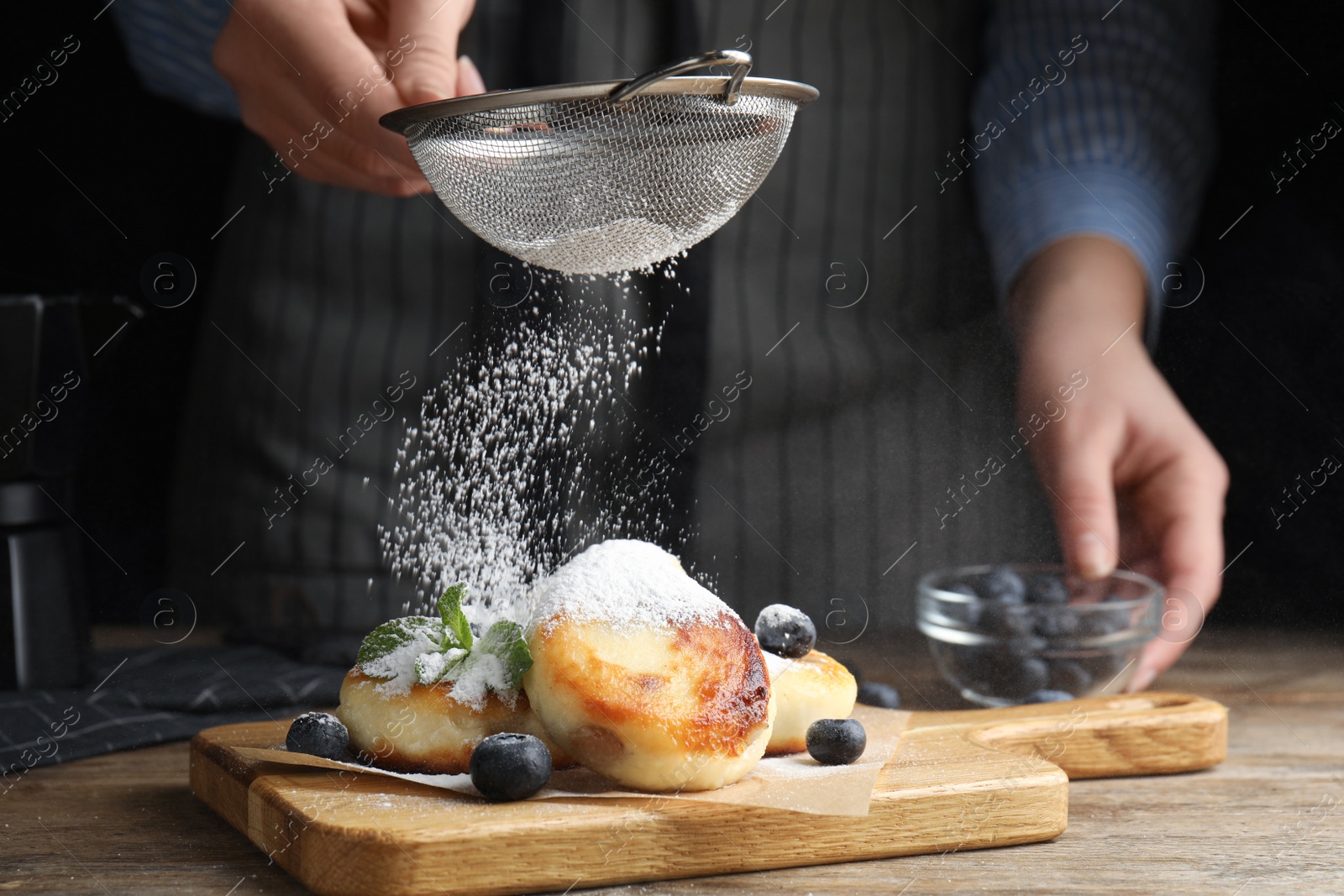 Photo of Woman sieving sugar powder on cottage cheese pancakes at wooden table, closeup