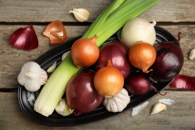 Plate with fresh onion bulbs, leek and garlic on wooden table, flat lay