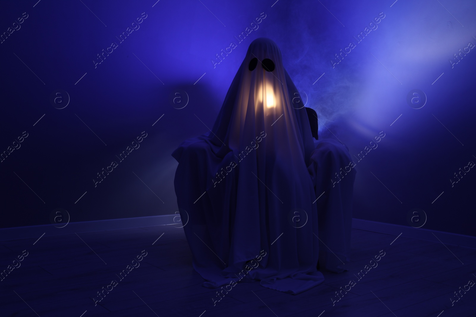 Photo of Creepy ghost. Woman covered with sheet sitting in armchair in blue light